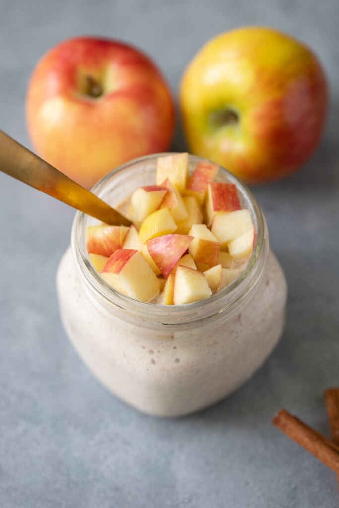 A jar of apple overnight oats with a gold spoon in it and two apples in the background