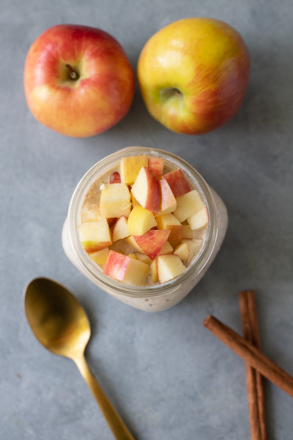 An overhead shot of a jar of apple overnight oats with apples, gold spoons and cinnamon sticks on the side