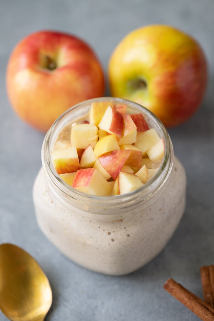 A jar of apple pie overnight oats with apples in the background and gold spoons on the side