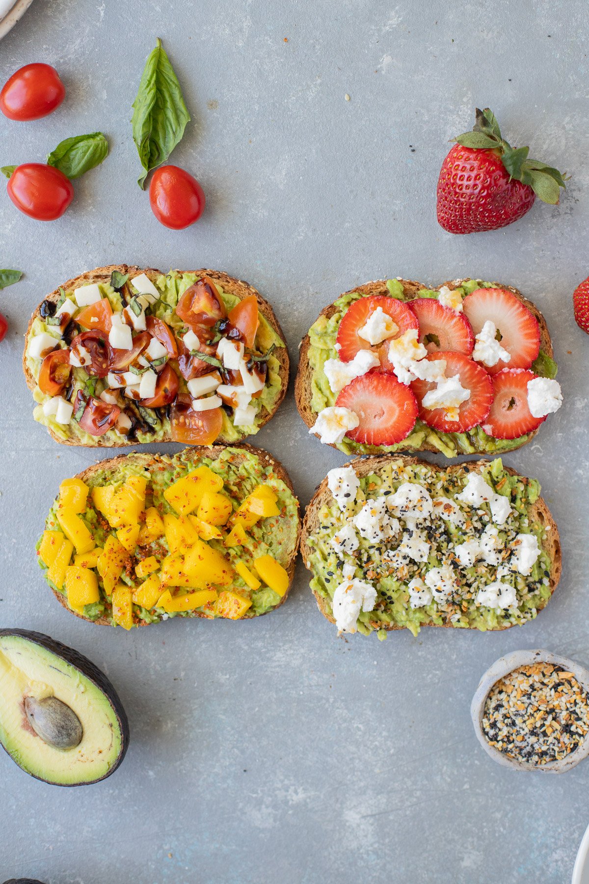 4 pieces of avocado toast (caprese toast, strawberry toast, mango tajin toast, goat cheese toast) on a grey background with tomatoes, basil, strawberries, avocado, and everything bagel seasoning on the side