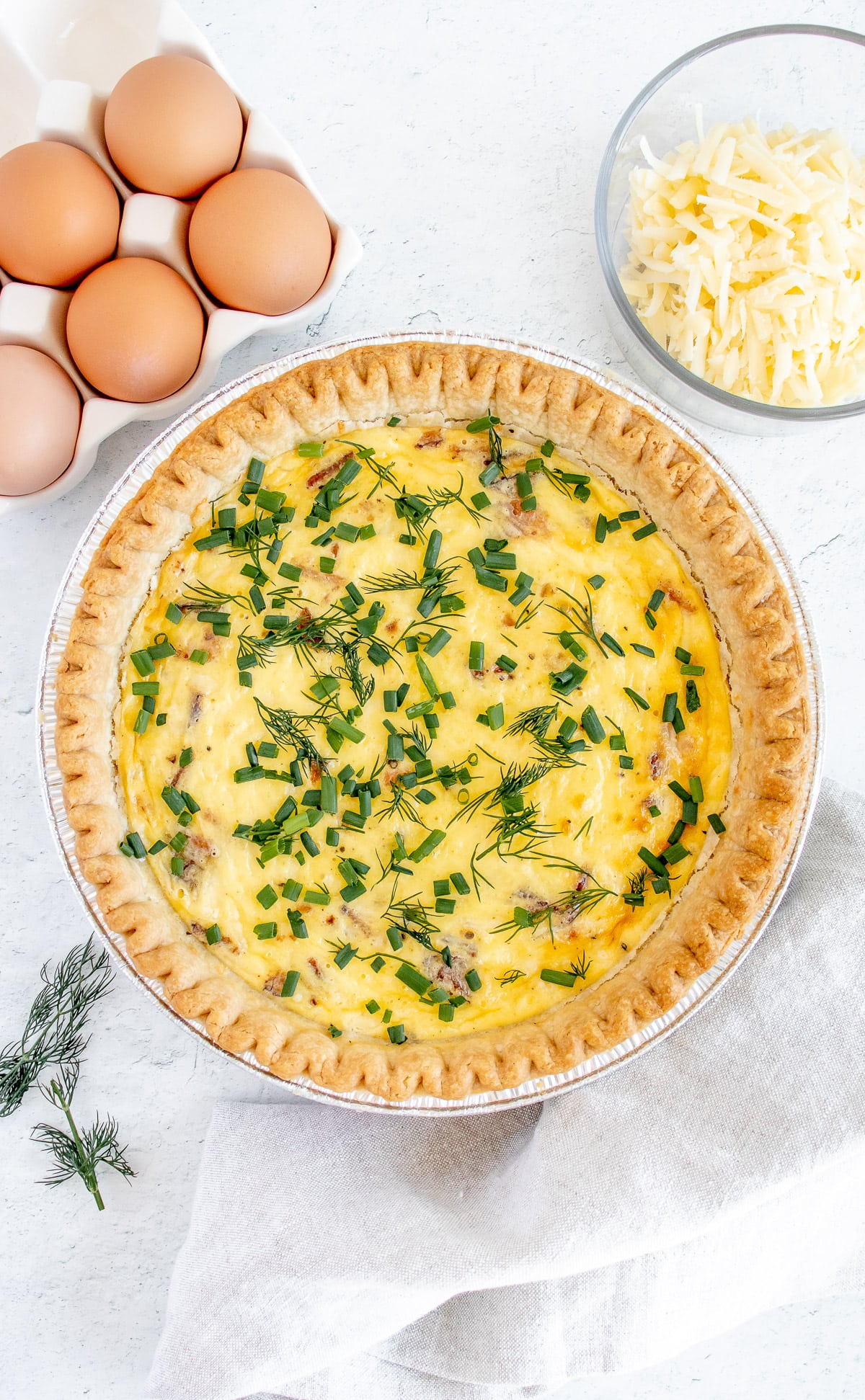 an overhead shot of bacon quiche with fresh herbs on top with eggs and cheese on the side on a white background