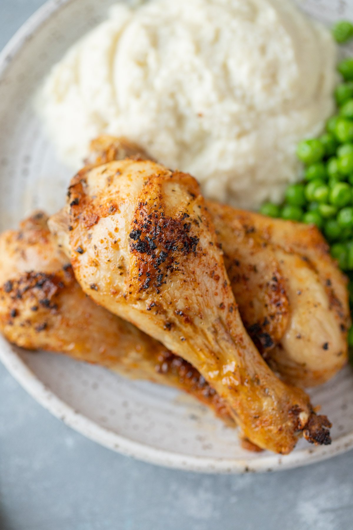 3 Baked Chicken Legs piled on a white plate with mashed potatoes and peas on the side on a grey background
