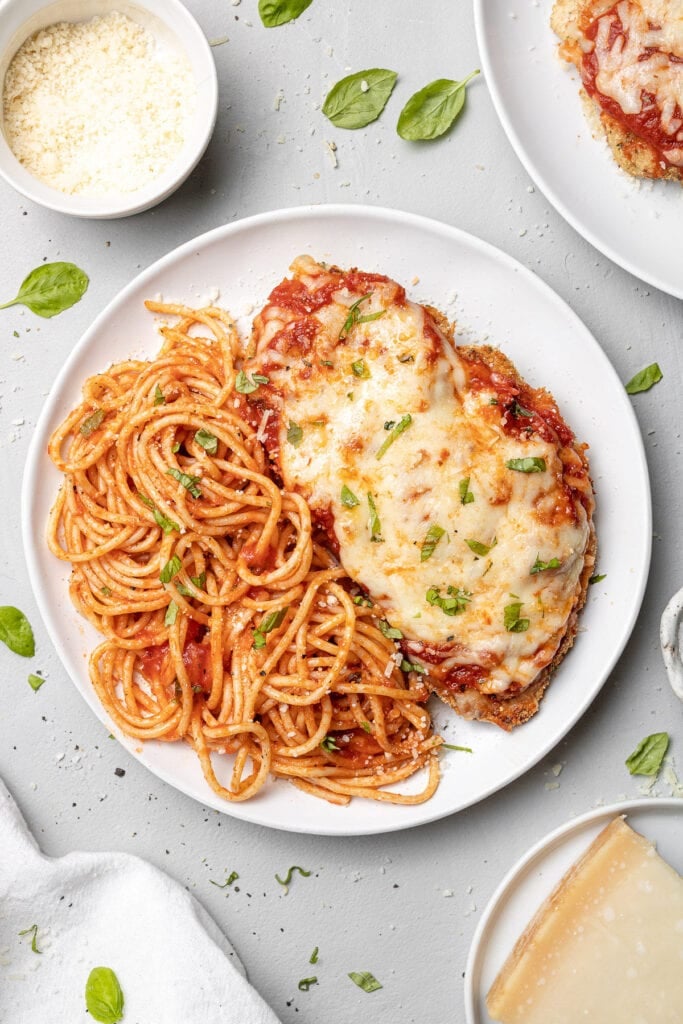 A plate of baked chicken parmesan with pasta and a bowl of cheese on the side