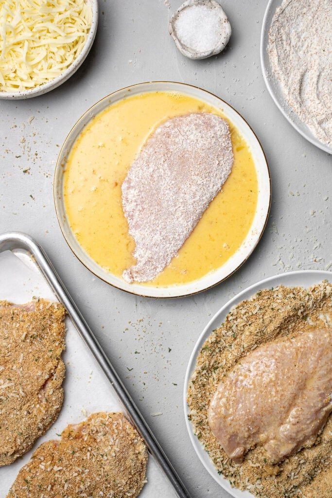 A chicken cutlet being dipped in a bowl of whisked eggs and a chicken cutlet being dipped in breadcrumbs with a sheet pan in the corner with breaded chicken cutlets on it for baked chicken parm