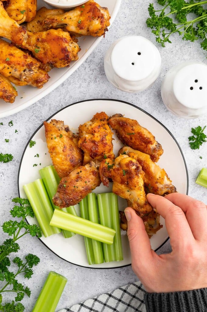 A hand grabbing a Baked Chicken Wing off a plate with celery on the side and a platter of the wings.