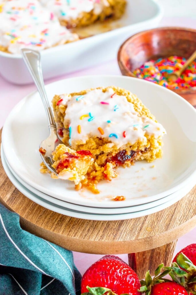 A piece of baked oats on a white plate with a bite on a fork resting on the plate with a baking dish of the baked oats in the background
