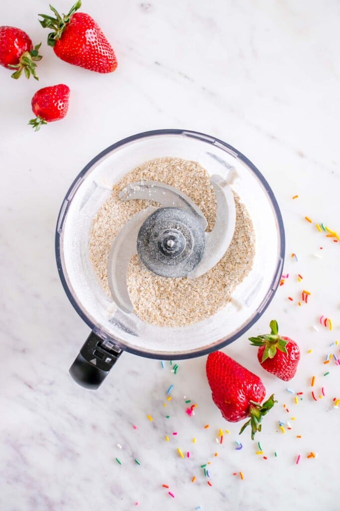 Oats in a food processor after being blended into oat flour with strawberries on the side