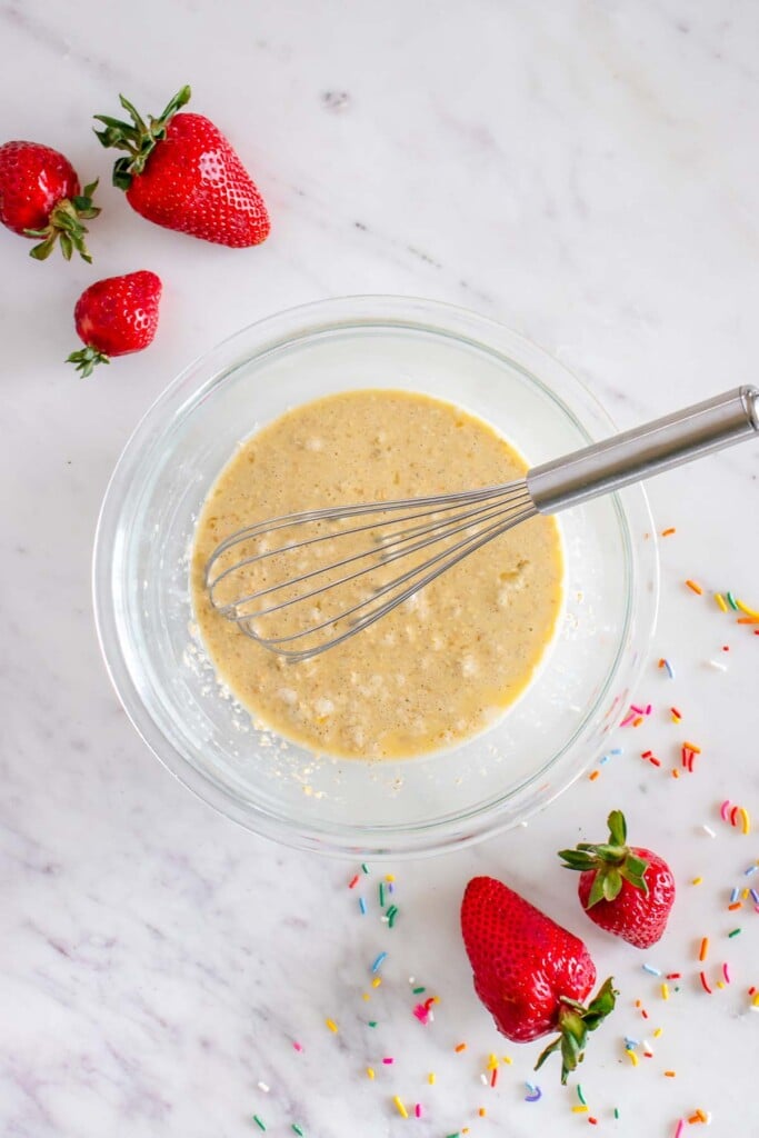 Batter for baked oats in a clear bowl with a whisk in it with strawberries on the side