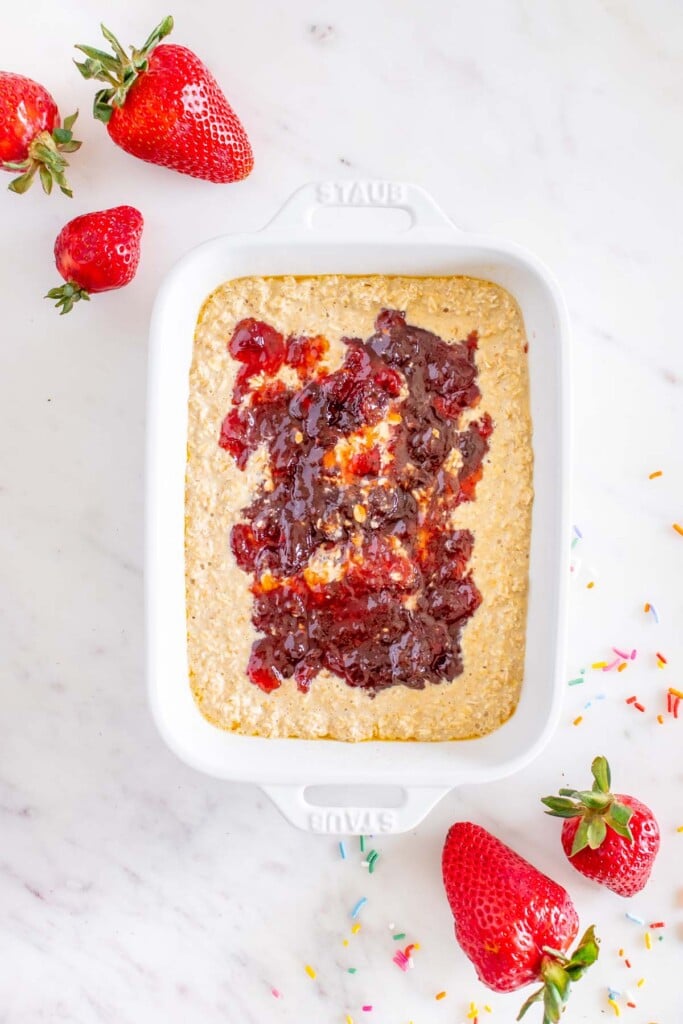 uncooked baked oats in a white baking dish topped with strawberry jam with strawberries on the side