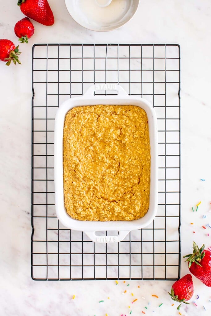 Fresh cooked baked oats in a white baking dish on a wire rack on a white background with strawberries on the side