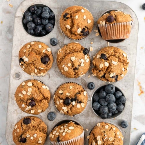 An overhead shot of a muffin tin with 10 banana blueberry muffins with blueberries around them on a white background