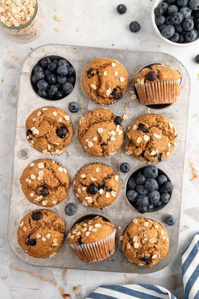 An overhead shot of a muffin tin with 10 banana blueberry muffins with blueberries around them on a white background