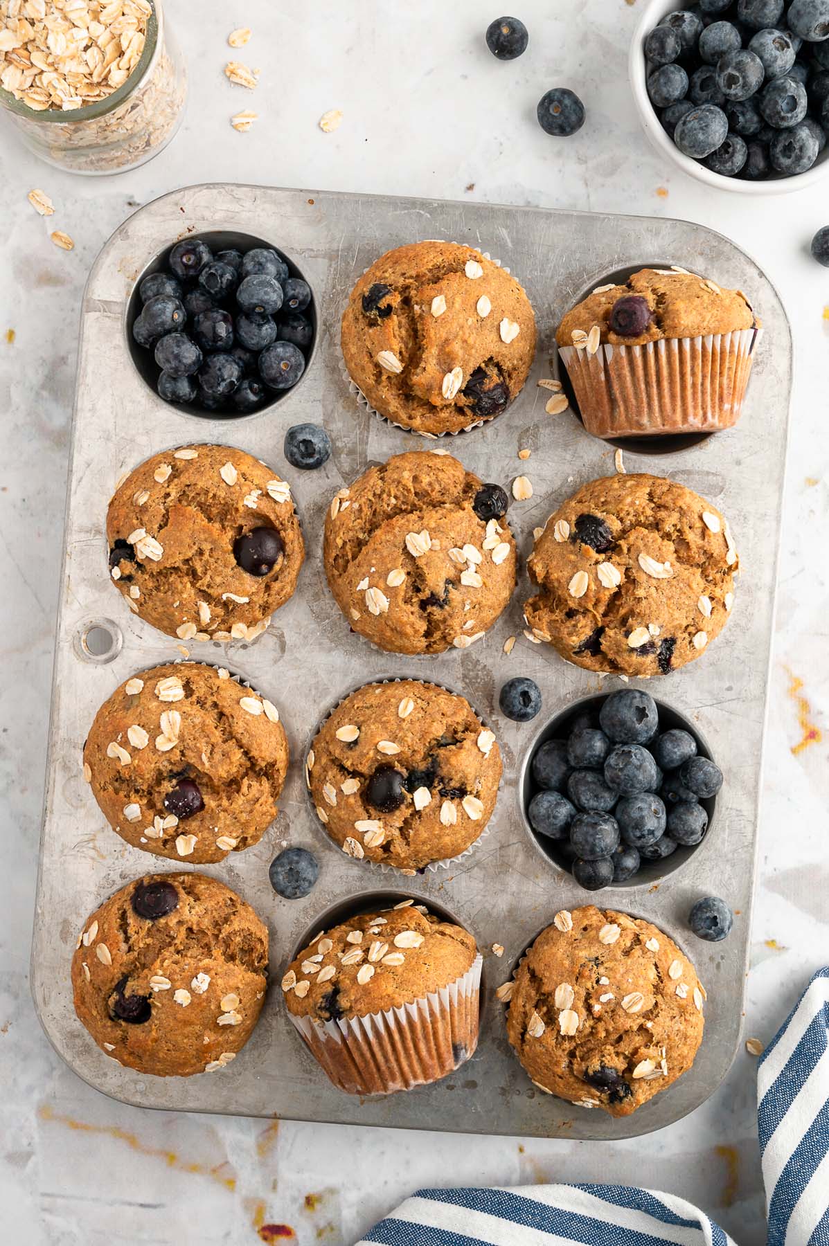 An overhead shot of a muffin tin with 10 banana blueberry muffins with blueberries around them on a white background