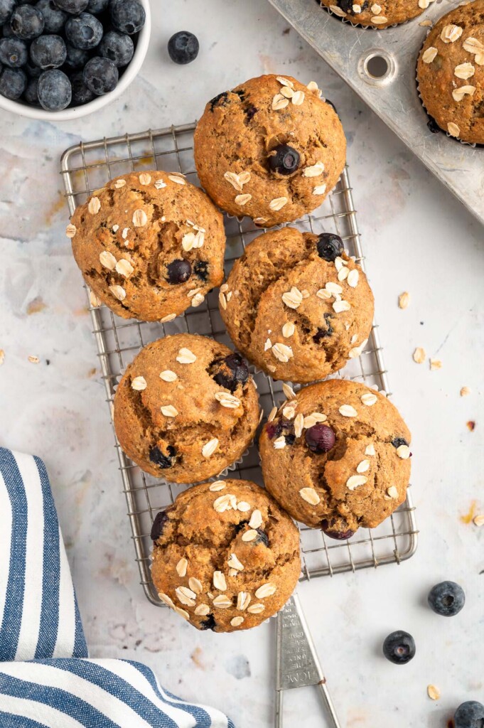 An overhead shot of 6 blueberry banana muffins on a cooling rack with blueberries on the side with the muffin tin on the side on a white background