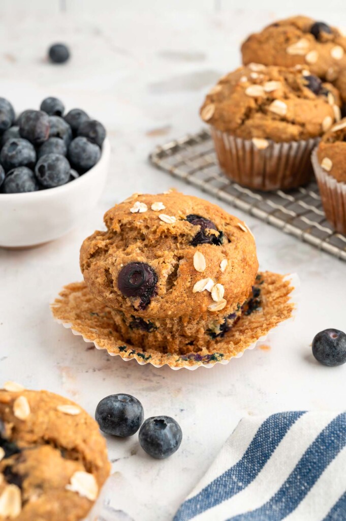 A single banana blueberry muffin with the liner pulled away and more muffins and blueberries surrounding it and some muffins on a cooling rack