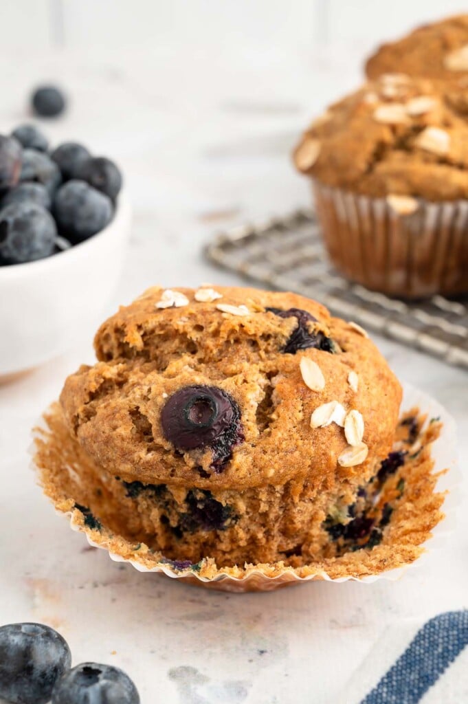 A single banana blueberry muffin with the liner pulled away and blueberries surrounding it and some muffins on a cooling rack in the background