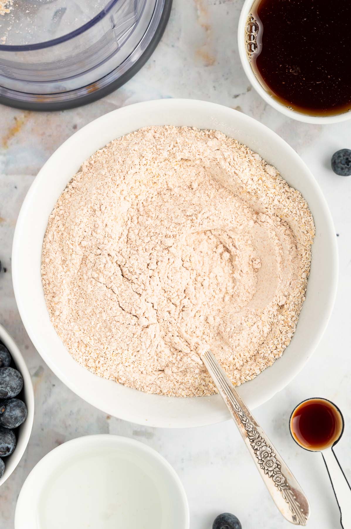 An overhead of a white bowl of dry ingredients (oat flour, whole wheat flour, baking soda, baking powder, and salt) after being mixed with a silver spoon for blueberry banana muffins with more ingredients surrounding it in white bowls