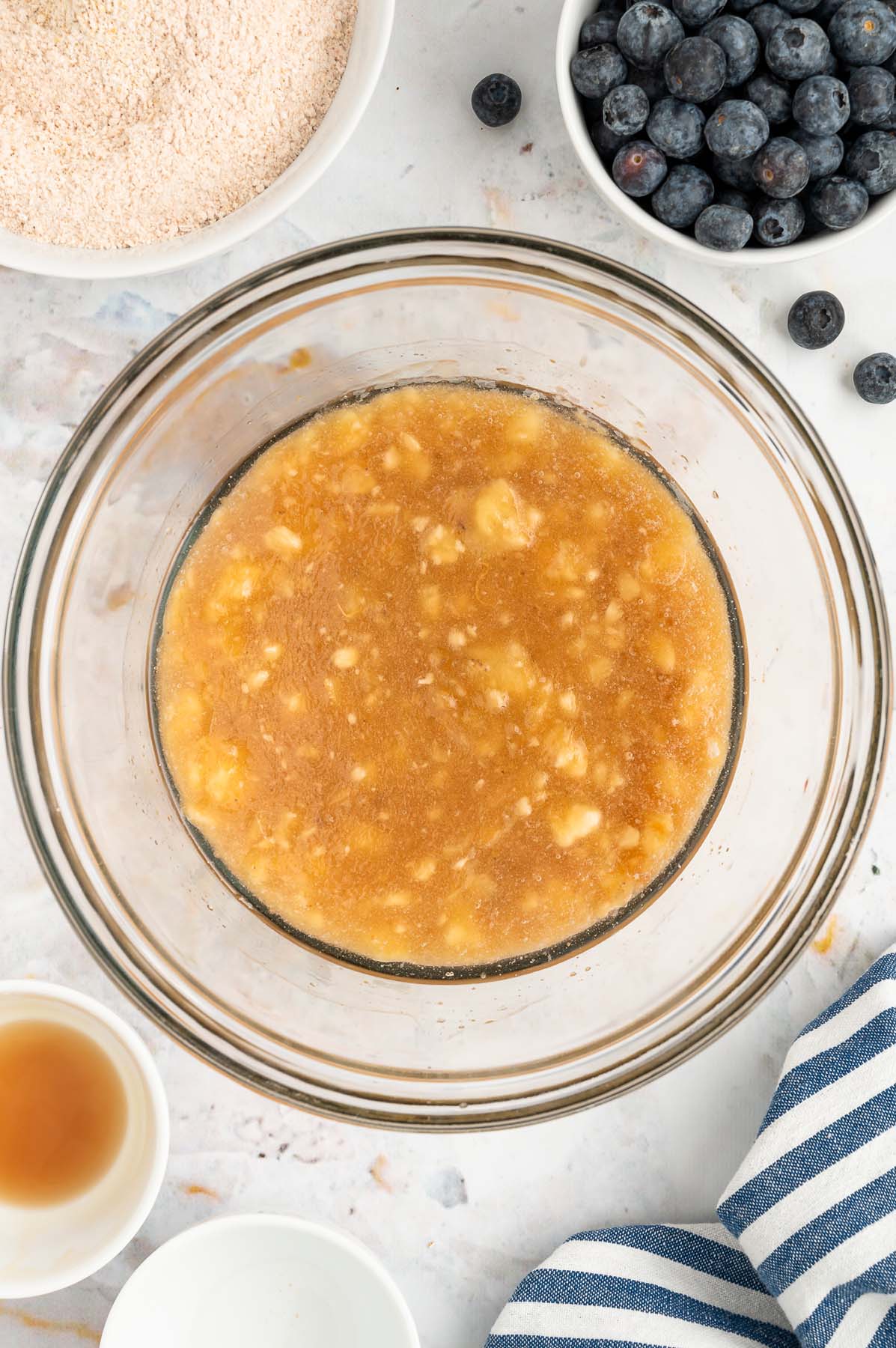 An overhead of wet ingredients (mashed bananas, eggs, maple syrup, coconut oil, and vanilla) for blueberry banana muffins in a clear bowl after mixing with dry ingredients and blueberries in bowls on the side on a white background