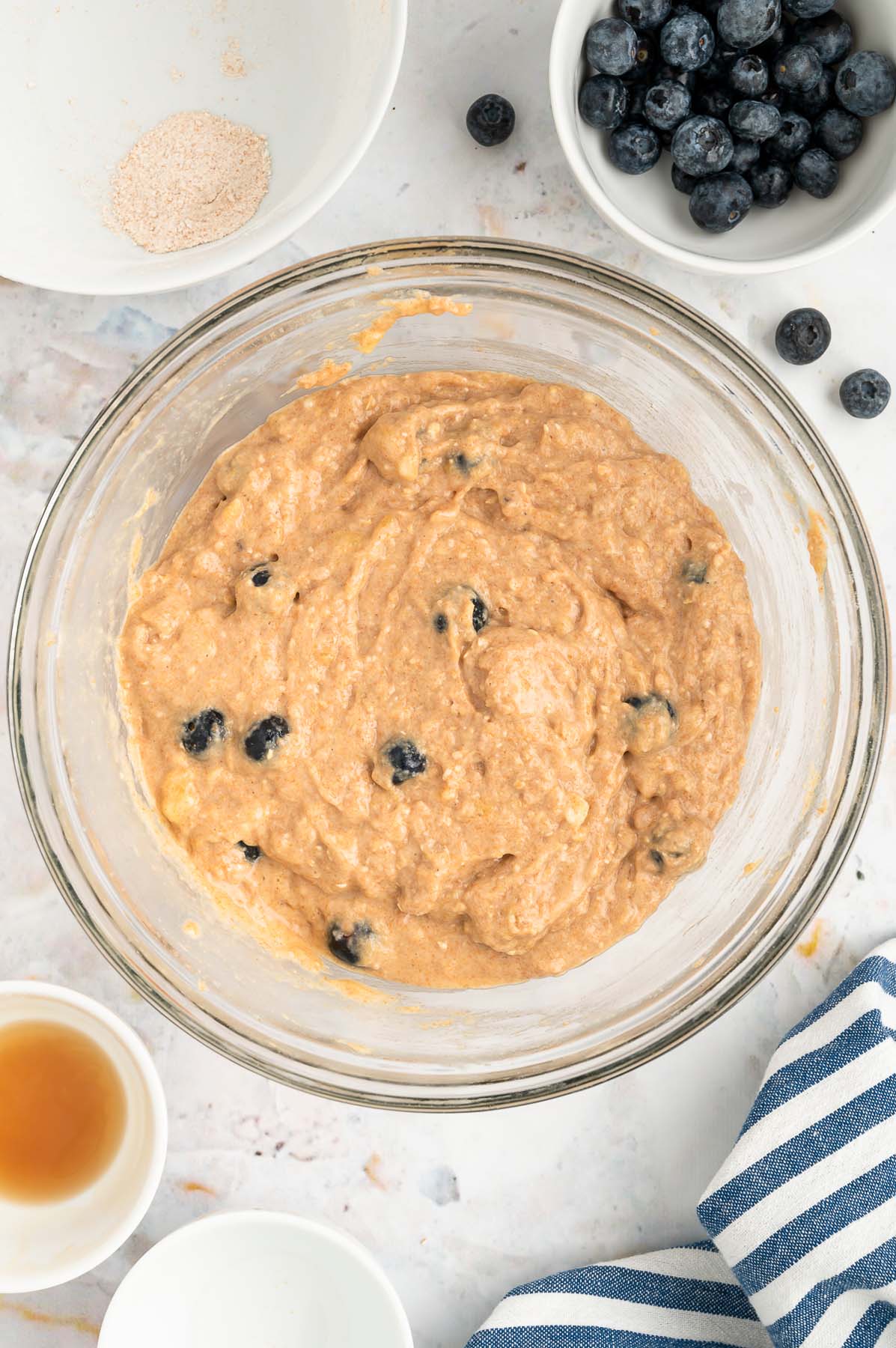An overhead of a clear bowl with blueberry banana muffin batter in it with a white bowl of blueberries on the side on a white background