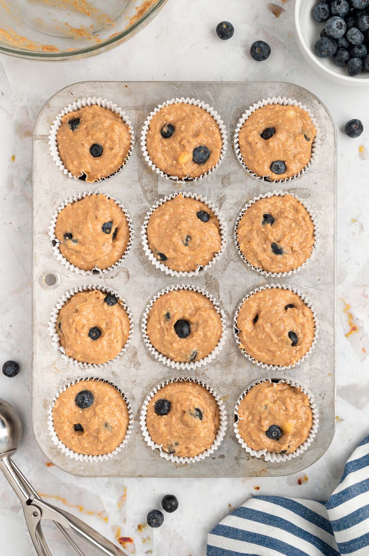 An overhead of a lined muffin tin with blueberry banana muffin batter in each of the 12 muffin spots on a white background