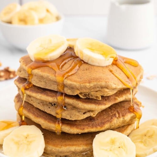 A stack of healthy banana pancakes on a white plate garnished with sliced bananas and maple syrup on top with a bowl of bananas in the background