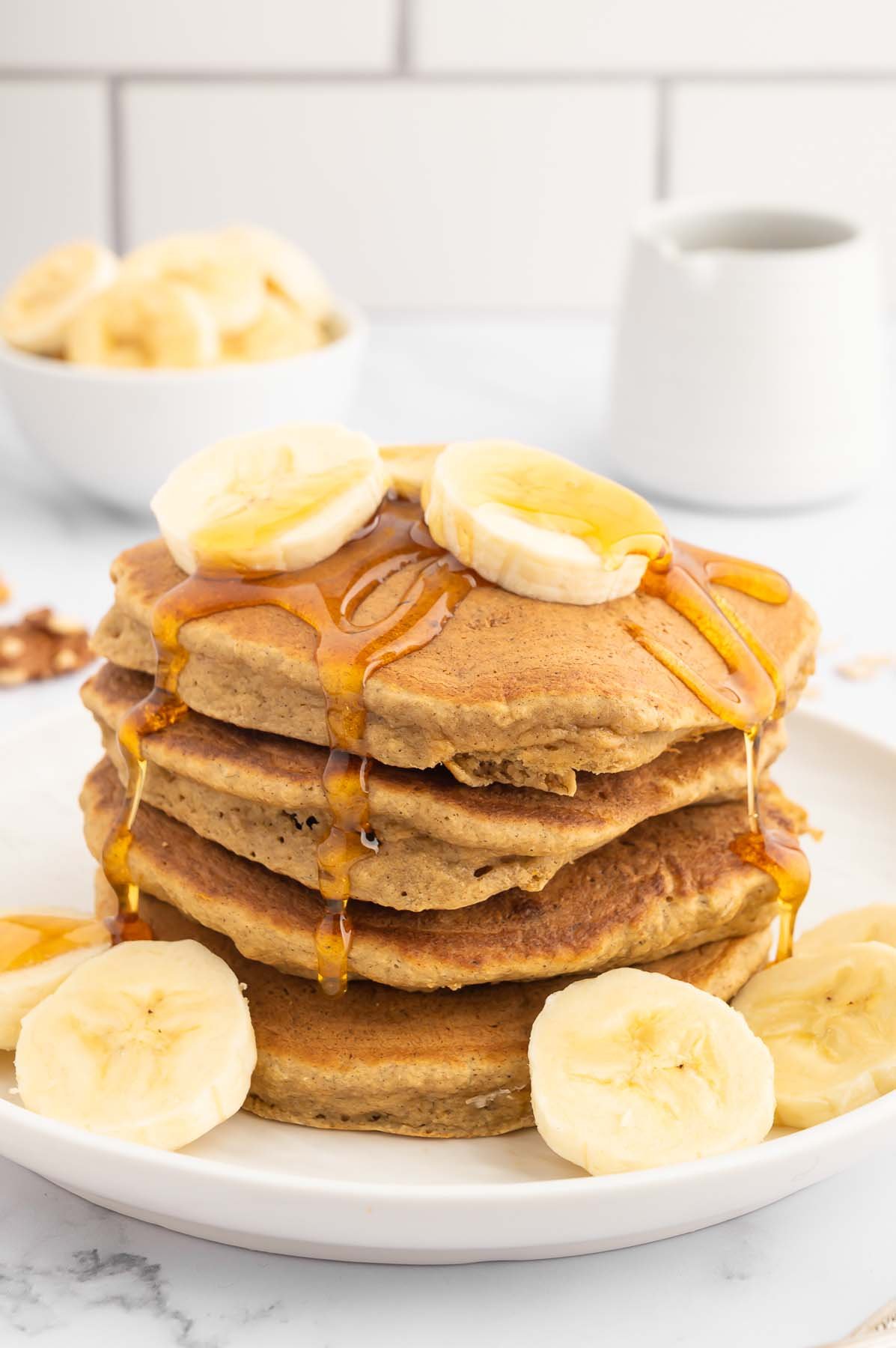 A stack of healthy banana pancakes on a white plate garnished with sliced bananas and maple syrup on top with a bowl of bananas in the background