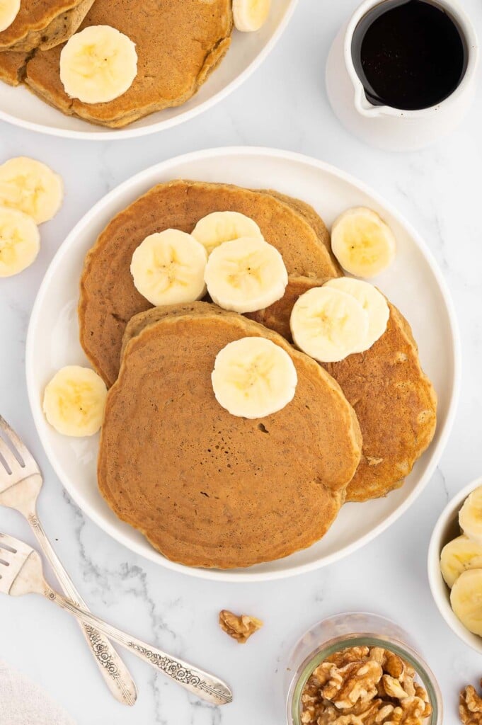 An overhead shot of banana pancakes on a white plate garnished with sliced bananas and a small white pitcher maple syrup on the side on a marble counter
