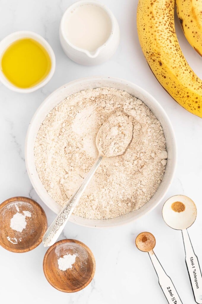 An overhead shot of dry ingredients for healthy banana pancakes in a shallow white dish with a spoon in it and ripe bananas on the side
