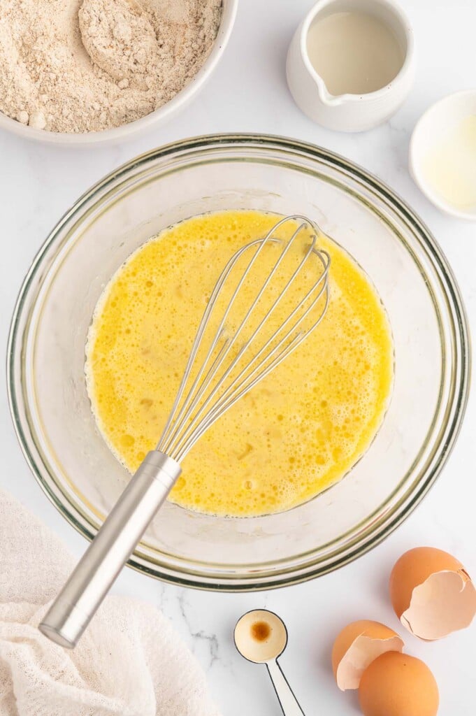 An overhead shot of wet ingredients for banana pancakes in a clear bowl with a whisk in the bowl on a marble counter