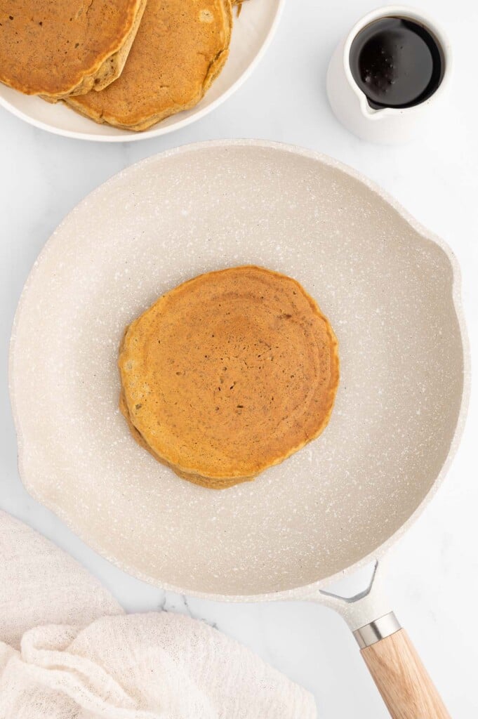 An overhead shot of a healthy banana pancake in a beige nonskillet skillet with a white plate of more pancakes and a small pitcher of maple syrup on the side