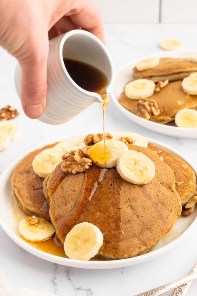 Three healthy banana pancakes on a white plate garnished with sliced bananas and a hand pouring maple syrup over them from a white small container another plate of pancakes is in the background