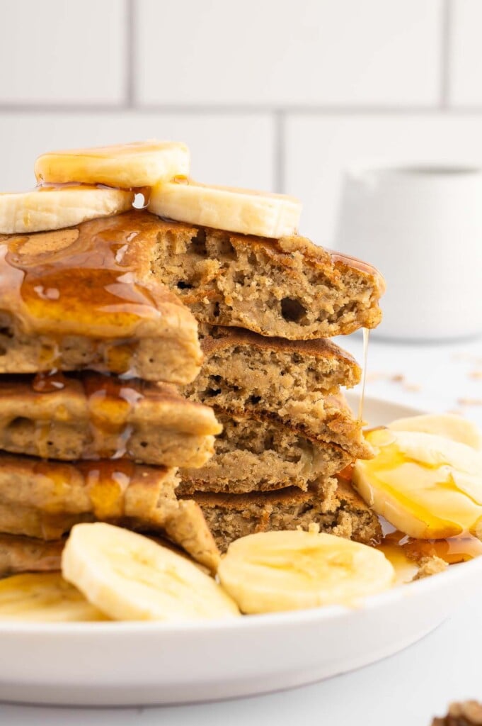 A stack of banana pancakes with a piece taken out they are garnished with sliced bananas and maple syrup on a white plate
