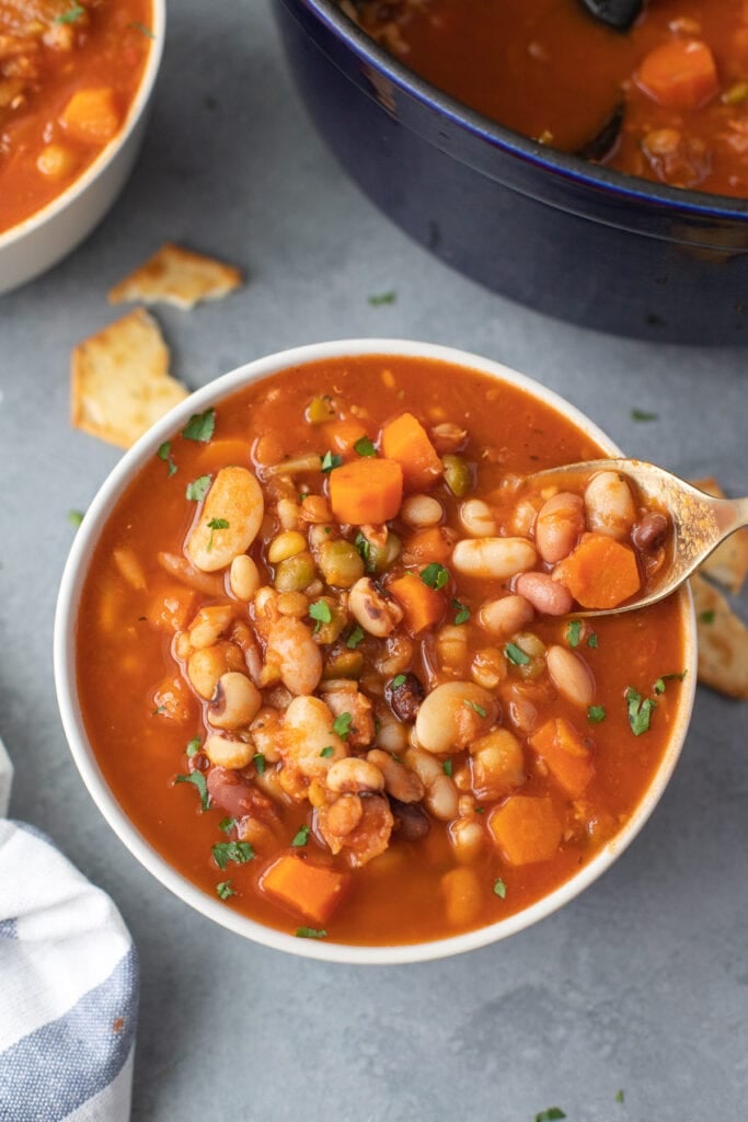 a white bowl of 15 bean soup with a spoon taking a bite out of it with a dutch oven (large pot) and bowl of soup in the background