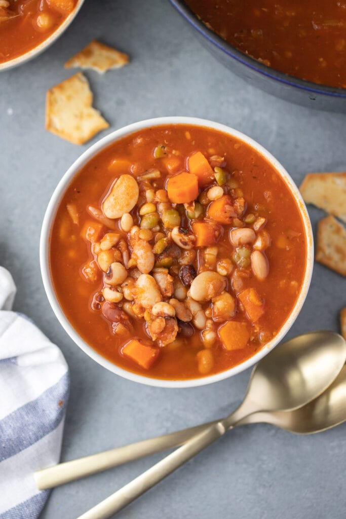 an overhead shot of a bowl of 15 bean soup with spoons on the side on a grey background