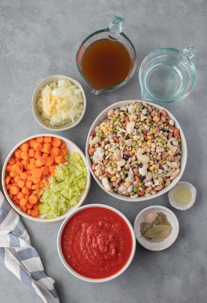 Ingredients for 15 bean soup in white bowls on a grey background: olive oil, onion, celery, carrots, salt, pepper, bay leaf, dried basil, dried oregano, dried parsley, crushed tomatoes, broth (veggie or chicken broth), water, 15 bean soup mix 