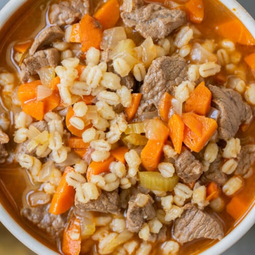 An overhead shot of a bowl of beef barley soup on a grey background with gold spoons on the side