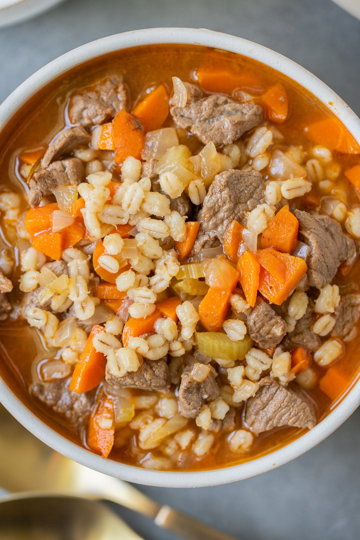 An overhead shot of a bowl of beef barley soup on a grey background with gold spoons on the side