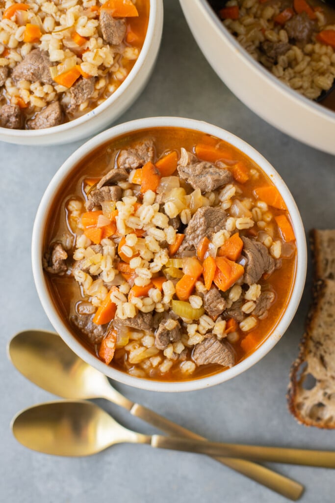 An overhead shot of a white bowl of beef barley soup with another bowl and pot of soup and gold spoons on the side