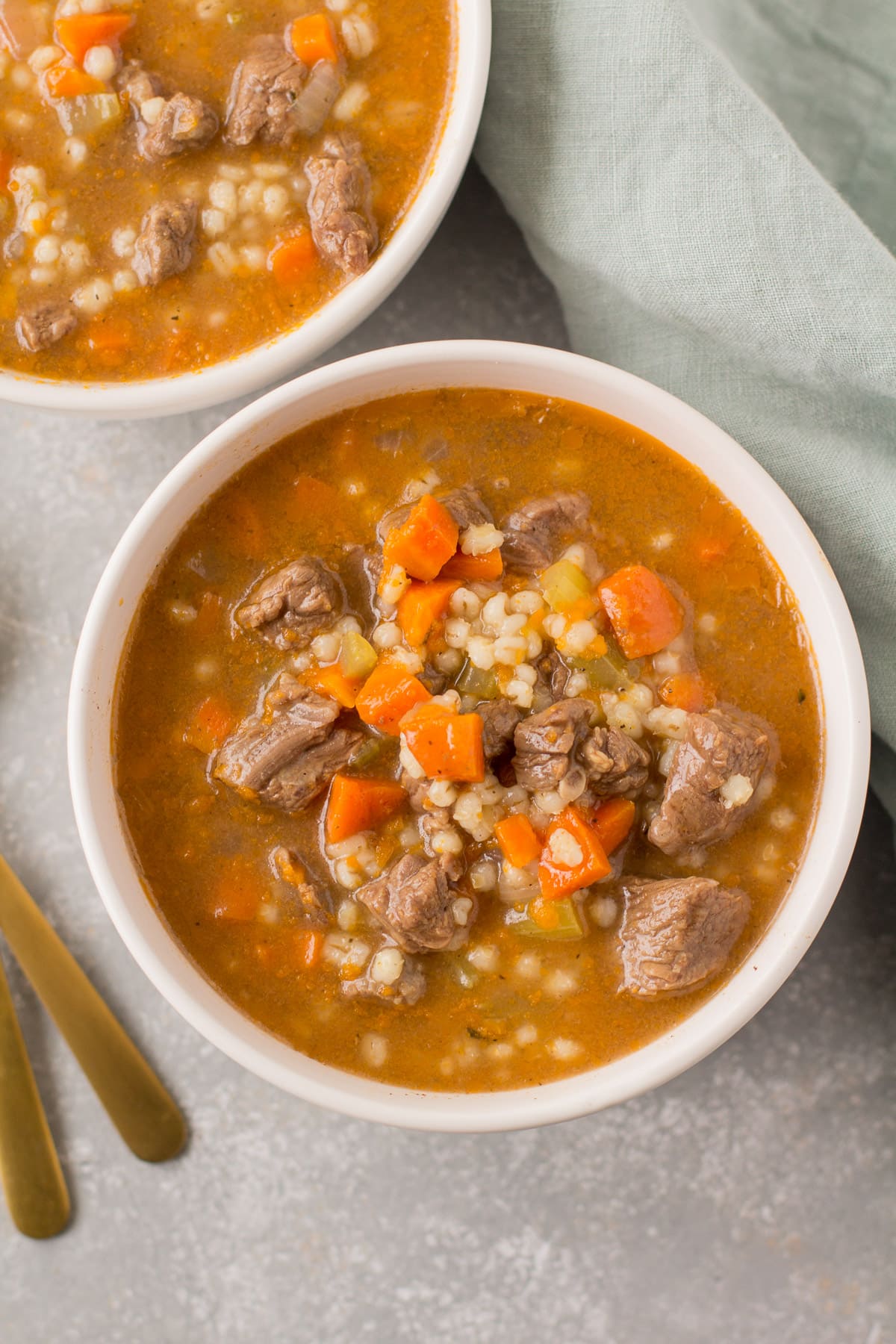 Healthy Beef Barley Soup in white bowls with a green linen and gold spoons off to the side.