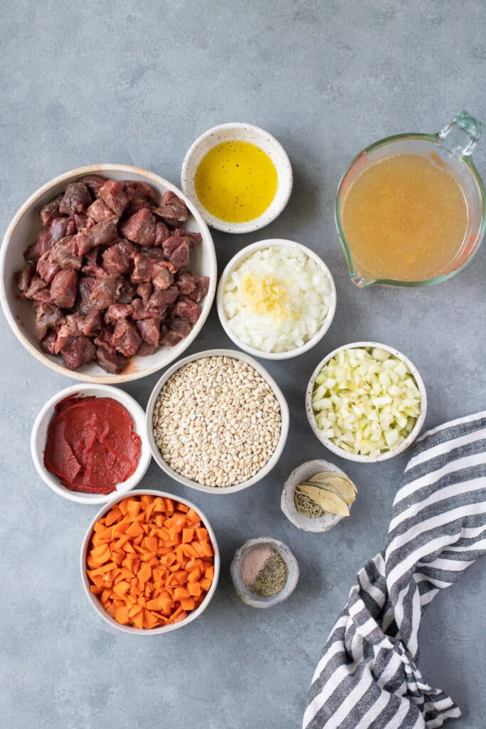 Ingredients for beef barley soup in white bowls: stew meat, olive oil, salt, pepper, garlic powder, rosemary, bay leaves, beef broth, pearl barley, veggies, tomato paste