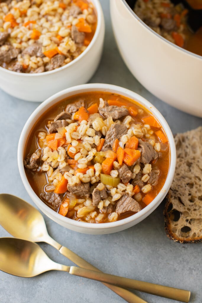 A bowl of beef barley soup with another bowl and pot in the background and gold spoons