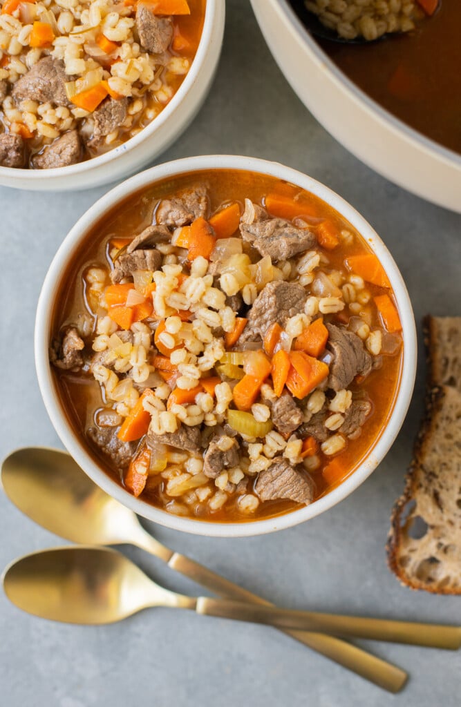 An overhead shot of a white bowl of beef barley soup with another bowl and pot of soup and gold spoons on the side