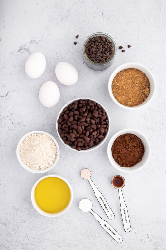 Ingredients for black bean brownies in small bowls: eggs, coconut sugar, black beans, oat flour, avocado oil, unsweetened cocoa powder, vanilla extract, baking powder, sea salt, semi-sweet mini chocolate chips