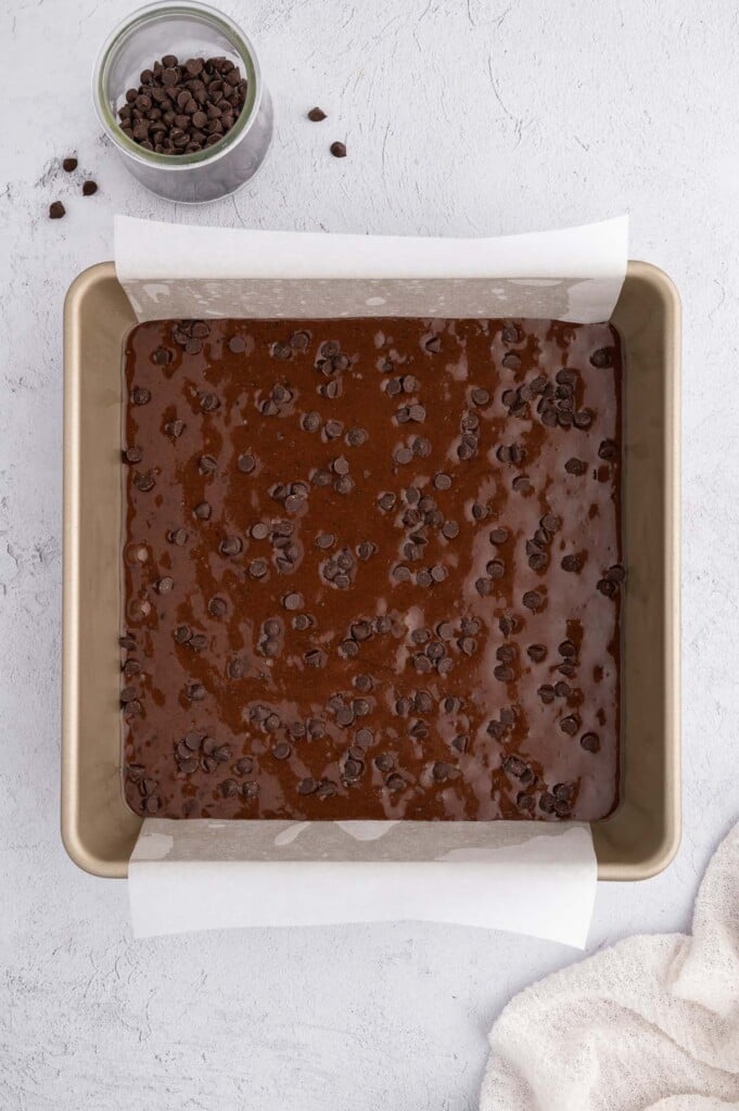 Black bean brownie batter topped with mini chocolate chips in a parchment lined baking pan with more chocolate chips on the side 