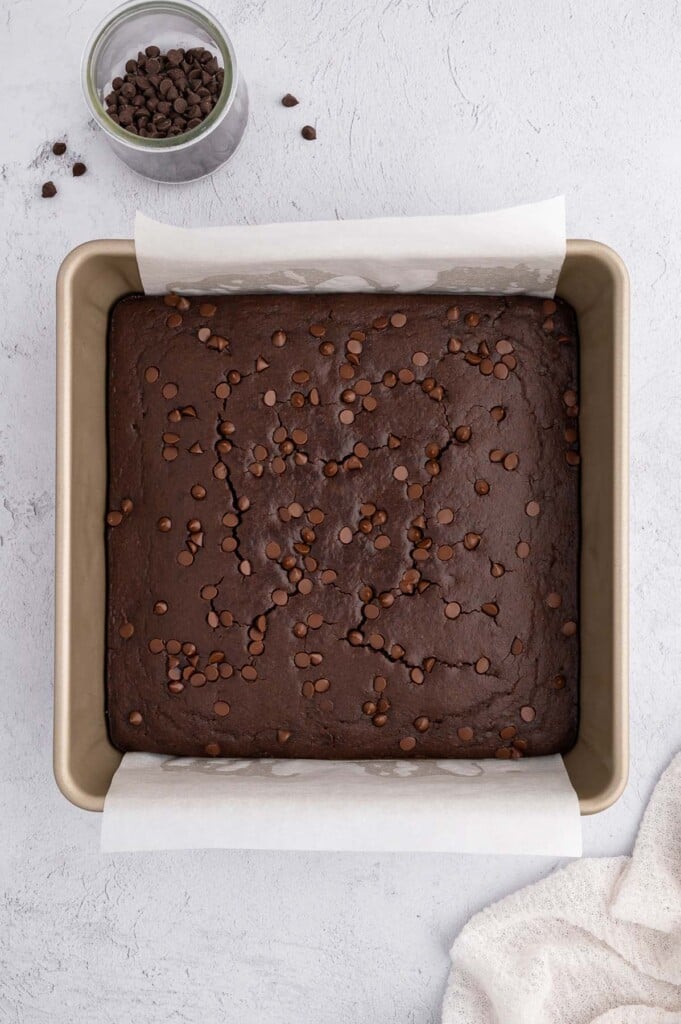 An overhead shot of black bean brownies right out of the oven in a parchment lined baking pan with mini chocolate chips on the side