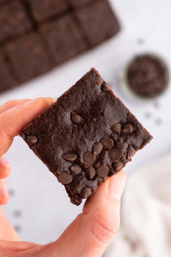 A hand holding a healthy black bean brownie with a pan of brownies and chocolate chips in the background