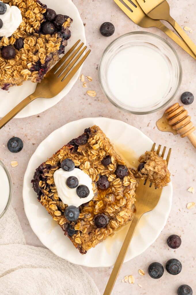 An overhead shot of blueberry baked oatmeal on a white plate with another plate, glass of milk, and berries off to the side.