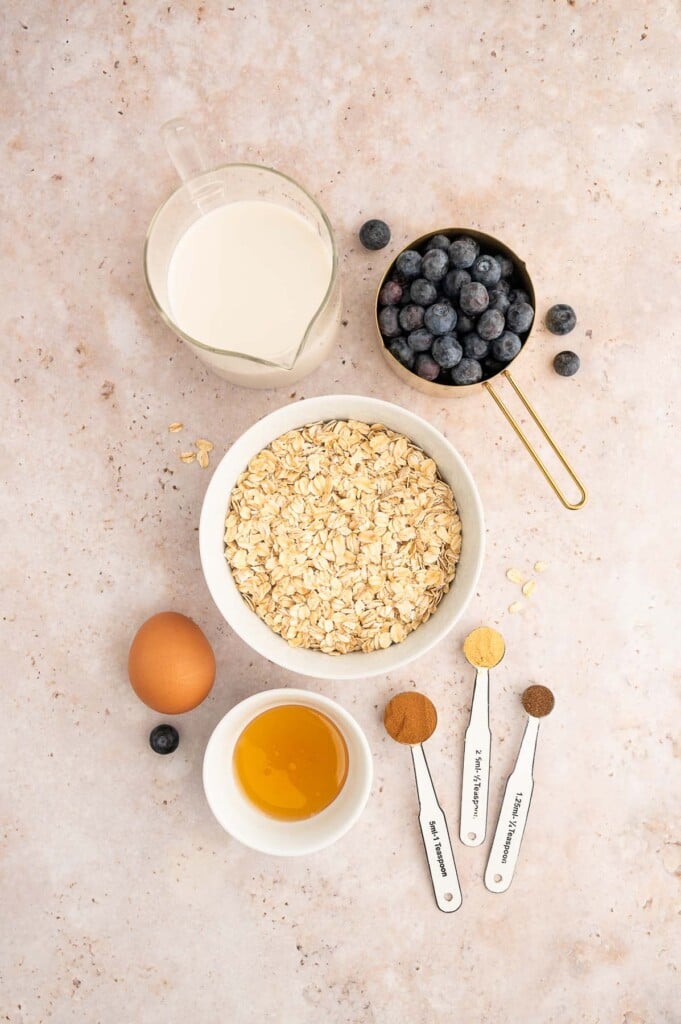 Ingredients for Blueberry Baked Oatmeal in small white bowls on a grey background: milk, cinnamon, ground ginger, nutmeg, old fashioned oats, blueberries, coconut oil, honey, egg