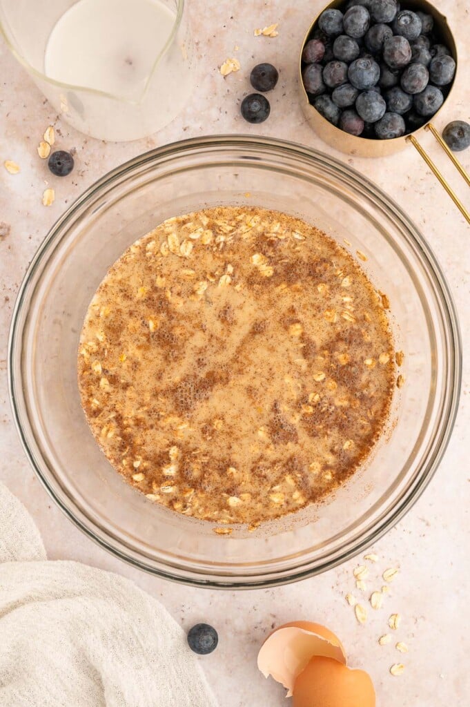 Wet and dry ingredients for blueberry baked oatmeal mixed in a clear bowl with blueberries on the side.