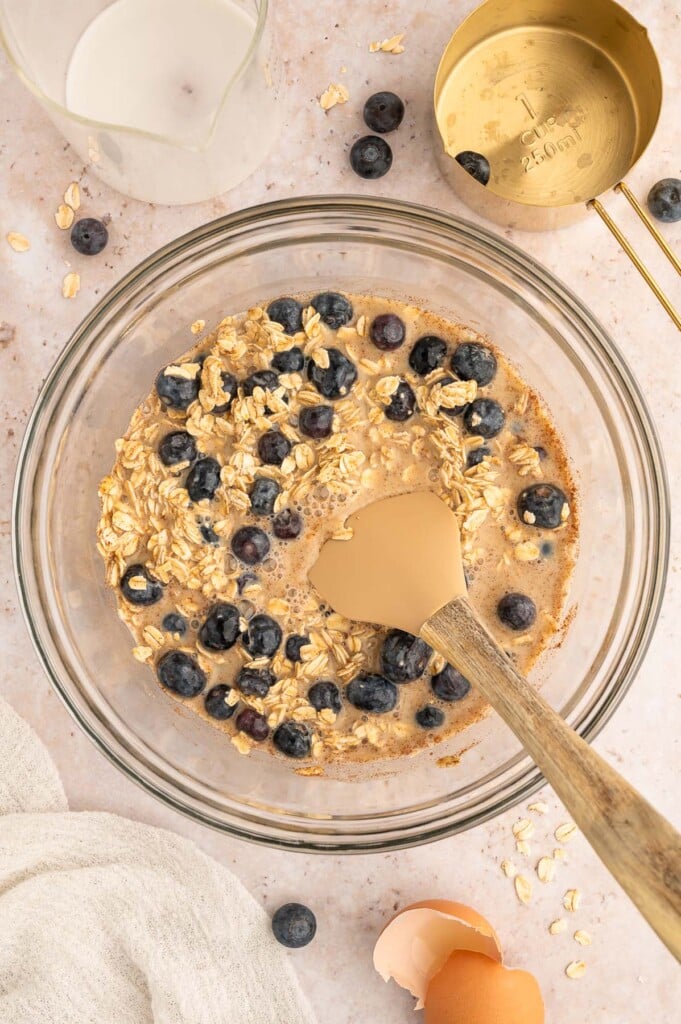Blueberries being folded into other ingredients for blueberry baked oatmeal in a clear bowl.